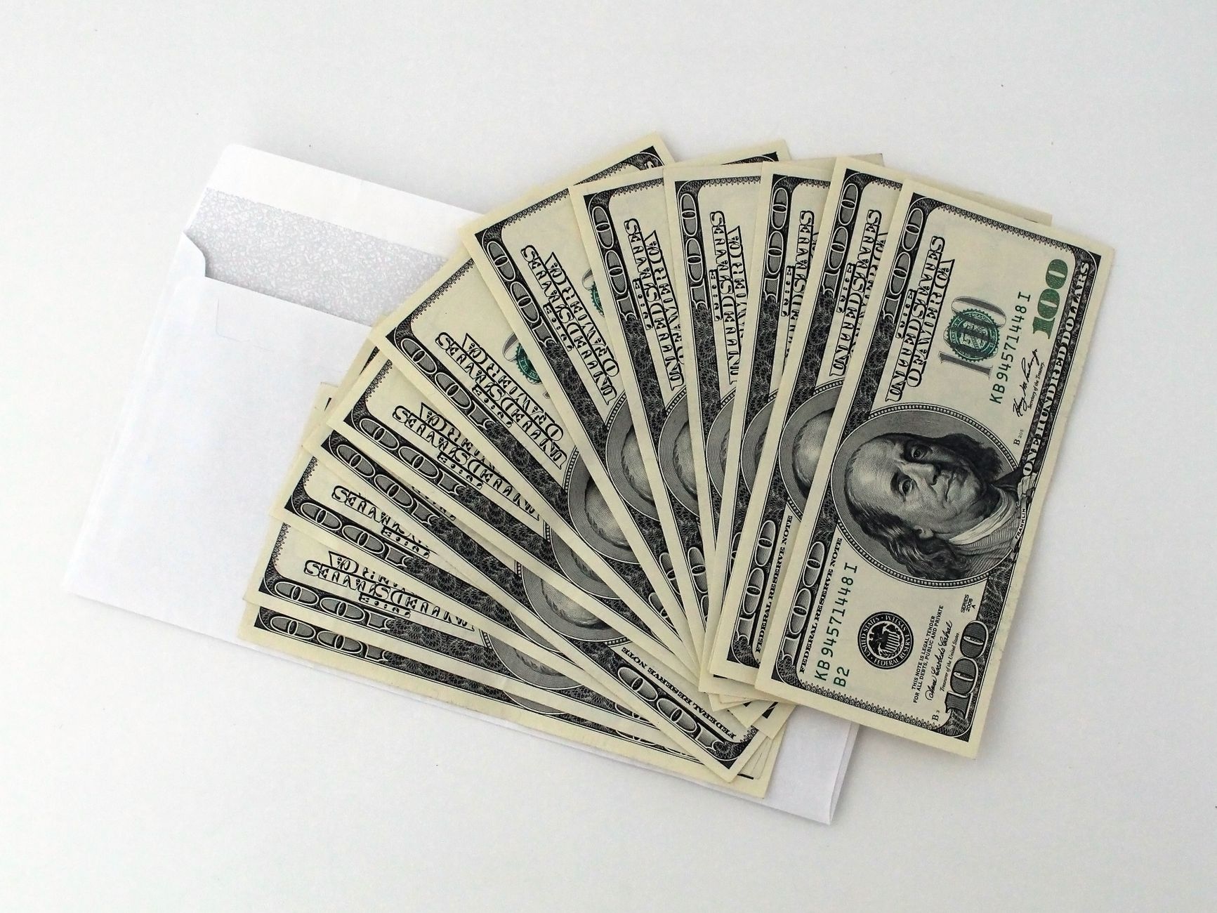 Abanico de billetes de US$100 parcialmente fuera de un sobre blanco sobre un fondo blanco.