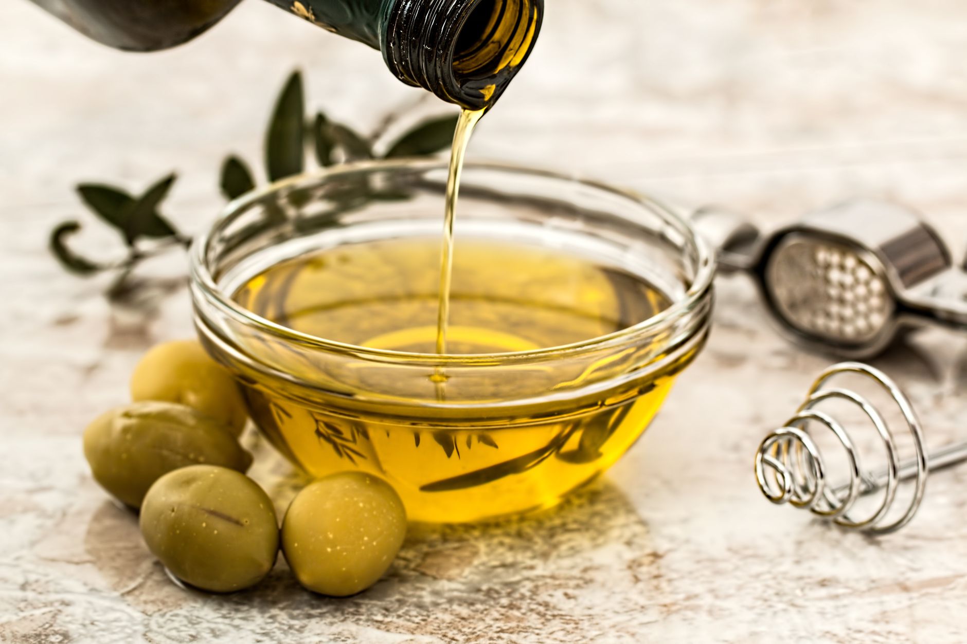 Primer plano de aceite de oliva siendo vertido en un tazón de cristal rodeado de aceitunas frescas y utensilios de cocina.